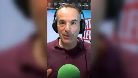 Martin Lewis is sat in front of a green mic with black headphones on. He is mid speech.