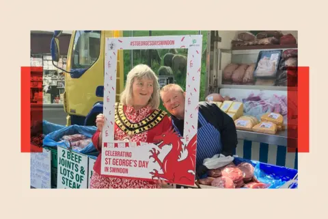Two women pose behind a blown up photo frame. One is a butcher working at a market stall and wears a stripey blue apron, the other wears a red dress and mayoral chain