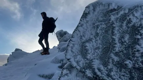 A walker, wearing crampons and with an ice axe, is on a high, snowy mountain path looking at the view. A rock which has ice on it is in the foreground.