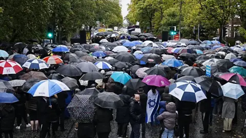 PA Media A large crowd of people, many holding umbrellas, stand in front of a stage flanked by stacks of large speakers. 