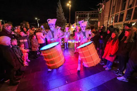 Sunderland City Council Two entertainers taking part in the community parade. They are both wearing white face make-up and dressed in white uniforms and hats. They are each beating a red drum as crowds look on.