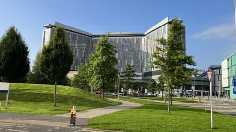 Queen Elizabeth University Hospital in Glasgow, a large modern hospital building with a curved glass façade, seen beyond a landscaped green lawn, trees and a pedestrian path on a clear sunny day.