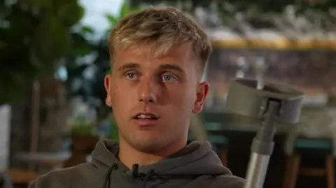 A young man with short blonde hair and blue eyes is speaking as he wears a grey hoodie. There is a grey and silver crutch next to him.