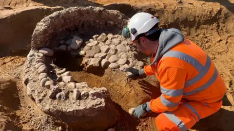 Oxford Cotswold Archaeology The back view of an archaeologist wearing an orange hi-vis jumpsuit and a hard hat. He is squatting by the remains of a medieval oven built of stone and mud. It has lost its roof and is emerging from yellow sandy soil.