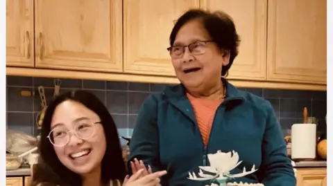 Two women, one is wearing clear frame glasses and sitting down and smiling. The elderly woman is standing also wearing glasses and smiling. They are in a kitchen and there are cupboards and kitchen role in the background