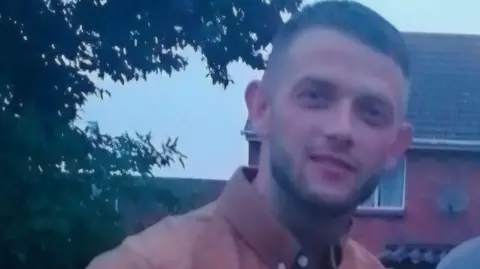 A slightly blurred photo of Danny who is smiling for the camera and wears a coral shirt. A house can be seen in the background.