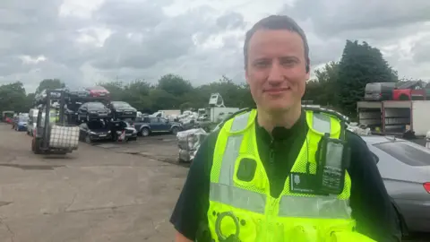 Charles Devereaux in the car scrapyard. He is wearing a high vis jacket and black top.