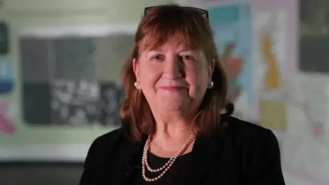 BBC Professor Dame Lorna Dawson, a mature lady with auburn hair, glasses on her head and wearing pearl earrings and a pearl necklace smiles at the camera. Behind her on the wall we see a map.