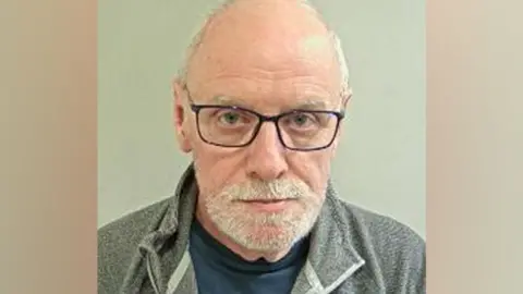 Macdonald's mugshot. He was very short white hair, balding on top nd a short white heard. He has black-framed glasses and is wearing a navy tshirt under a grey zipped top