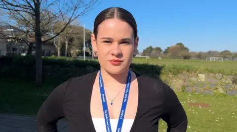 An 18-year-old student. She has slicked back dark brunette hair and three silver hoops in each earlobe. She is wearing a white top and a black cardigan.