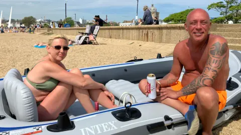 Henry Godfrey-Evans/BBC Carl Smith and Deanna Barrett sitting in a grey inflatable boat on the beach. Deanna is wearing a green bikini and Carl is wearing orange swimming shorts. Both are smiling on a sunny day.