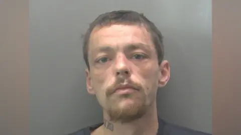Cambridgeshire Police A police mug shot of Ashley McMillan. He has light brown hair and a goatee beard. He is wearing a grey T-shirt. 