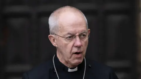 PA Media Justin Welby, the Archbishop of Canterbury, pictured wearing his cassock while standing in front of some large and dark wooden doors.