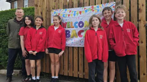Six students and the head teacher are standing in front of a sign that reads 'welcome back. 