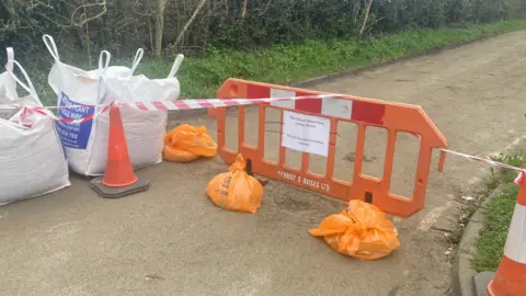 JUSTIN DEALEY/BBC An orange plastic barrier, about a car-wide, sacks of rubble, traffic cones and tape block off a narrow road. 