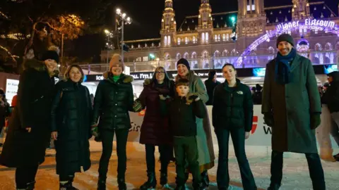 Vienna Tourist Board/Stefan Simonovic A group of eight residents from Dull ice skating in front of Vienna's town hall. They are wearing winter clothes and smiling at the camera. The town hall is lit up in the dark and there are purple lights in the background.