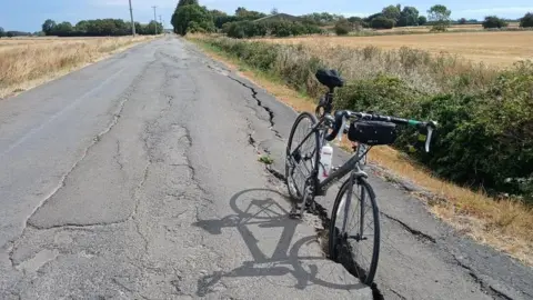 A black racing bicycle is standing inside a large crack in a rural road on a sunny day.