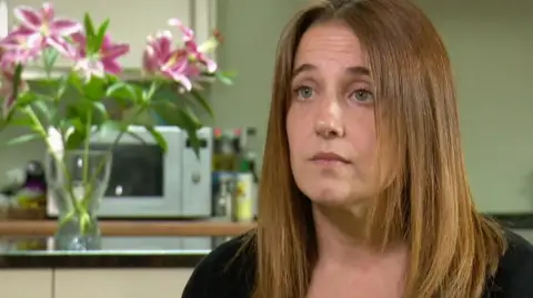 BBC Louise Slorance looks slightly off camera. She has shoulder length light brown hair and is wearing a black cardigan. She is sitting in front of a green wall in her kitchen. There is a vase of pink flowers on the island, which has a black worktop and white cupboards. In the background, there is a silver microwave underneath one cupboard on the worktop, next to a collection of bottles of oil.