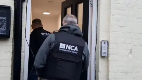 National Crime Agency The image shows two individuals wearing black tactical-style vests marked “NCA” and “National Crime Agency”. They appear to be entering a building through an open doorway. 