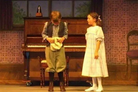 Supplied Two young actors are on stage in front of a piano. Bella Ramsey is on the right wearing a dress and a boy on the left has his head down and is holing a cap.