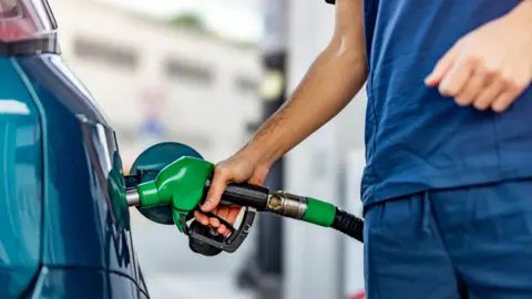 Getty Image Young man wearing blue overalls is refuelling his blue car at petrol station. The fuel hose in his hand is green. 