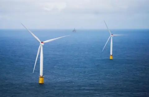 Two white wind turbines with yellow bases in a blue sea with a ship seen in the distance
