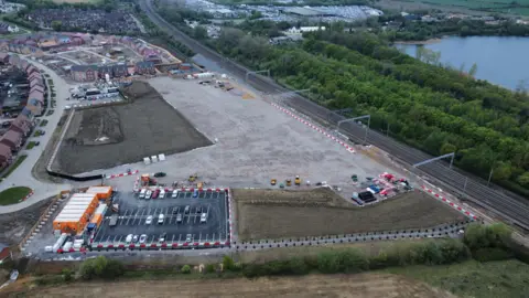 Ant Saddington/BBC Aerial view of a construction site with scattered equipment and vehicles, nearby are residential houses and a railway track.