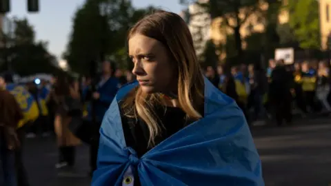 A woman stands looking to her side at a protest wrapped in a blue Ukraine flag.