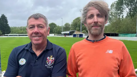 Glenn Eastick on the left is a man in his 50s with grey hair and his arms folded wearing a blue top. He is standing next to Ryan McKnight who has a lots of untidy hair and a beard and is wearing an orange top. Behind is a football pitch 