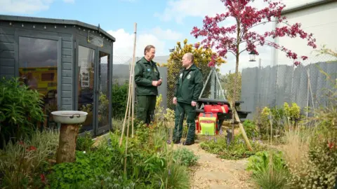 East of England Ambulance Charity Two ambulance crew members, both men, standing inside a garden with green shrubs and a pink-leaved tree and a grey outbuilding with glass windows.