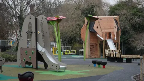A children's play area which includes two large slides designed to resemble elephants. One is grey with a small slide and the larger slide can be seen in the distance which has wooden cladding in the shape of an elephant's head, the slide acting as the trunk.