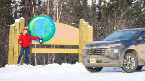 Ash Carter Ash Carter is standing in a red coat next to a large wooden road sign. He's gesturing to a large earth picture, with "Arctic Circle" written next to it. A black, dirty SUV is parked to the right and the ground is covered in snow.