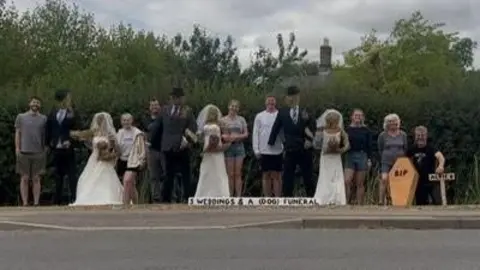 Jim Nelson A line-up of eight real people surrounding three scarecrow couples who are wearing wedding dresses and suits complete with black hats, at the far right is a small wooden coffin bearing the letters 'RIP'. A sign next to that says 'Alfie' who was the family dog that died last month. A longer white sign on the ground reads 'Three weddings and a (dog) funeral'.