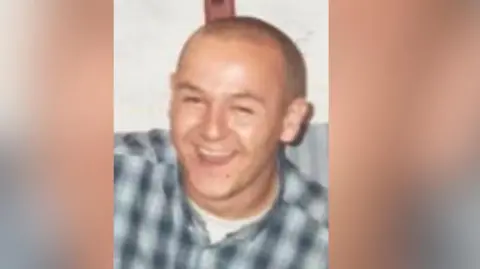 Merseyside Police Vincent Lawrence smiles as he is photographed. He is wearing a blue-and-white checked shirt, buttoned up over a white t-shirt.