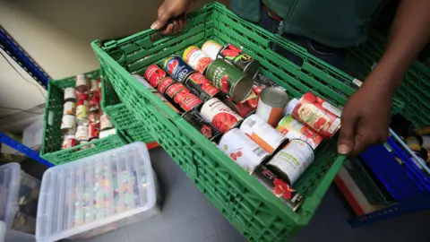 Crates on the floor full of tinned food. The floor is made of a blue tile. There are two hands holding a green crate filled with tinned chopped tomatoes. 