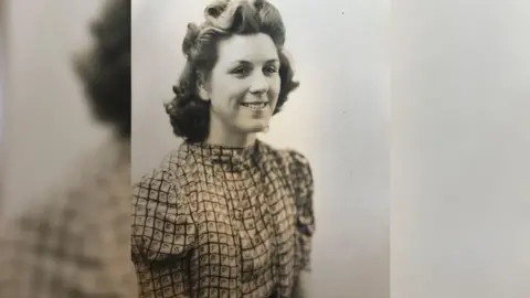 Hilda Bond A black and white photograph of Mrs Bond when she was younger. She is wearing a high neck blouse with a square pattern print on it. She is smiling and glancing at the camera with her hair down but styled into position