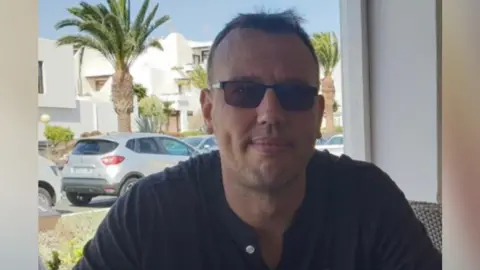 Sarah Healy Danny. A man with short brown hair who is sat outside at a table, wearing a blue top and glasses. 