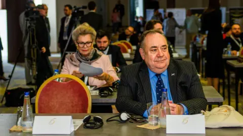 Academia de Diplomacia Cultural Alex Salmond sentado em uma mesa com uma placa branca à sua frente. A cadeira vermelha com moldura dourada ao lado dele está vazia. Salmond está vestindo um terno escuro com camisa azul e gravata azul escura, com um distintivo de lapela Saltire.