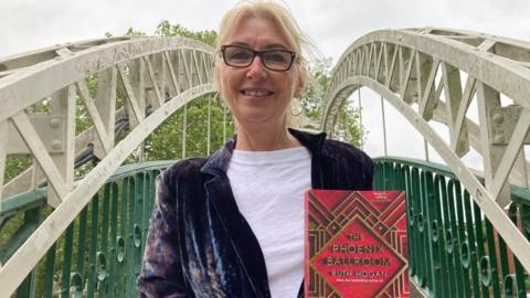 Ruth Hogan standing on The Suspension Bridge in Bedford holding her book