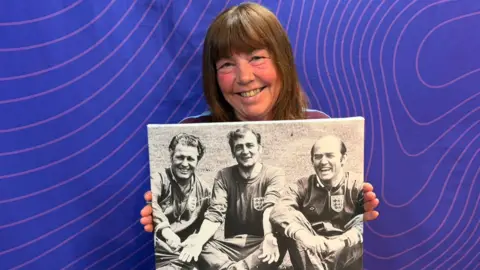 A woman with long brown hair holding up a large black and white photograph of three men in England football tracksuits