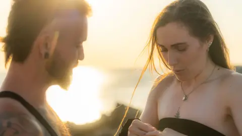 Ben Fiore Alain Branquet has a beard and is watching Jade Goracy look at the engagement ring. She is wearing a black bandeau top.