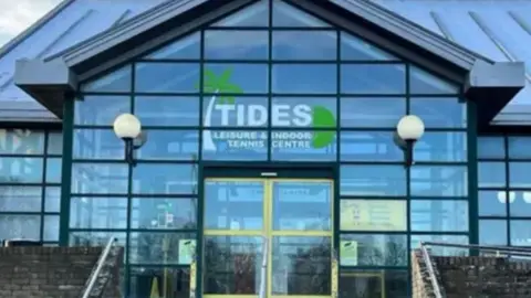 a glass-fronted building with a pitched roof, with Tides Leisure and Indoor Tennis Centre written above the door