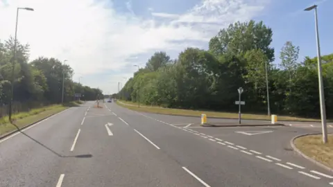 Google A straight road with two lanes and an extra turning section where it meets a T-junction, which is Bishops Rise. There are verges and trees on either side. 