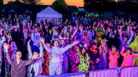 St Elizabeth Hospice Dozens of people wearing pyjamas and other brightly coloured costumes standing in the twilight with their arms held aloft.