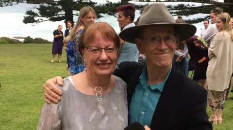 Geraldine Schirmer Geraldine has short hair and is wearing glasses. She is wearing a top with feathers on it. Donald is wearing a broad brimmed hat. He has glasses and a dark coloured suit jacket and blue shirt. They are at an event and other people stand near the couple.