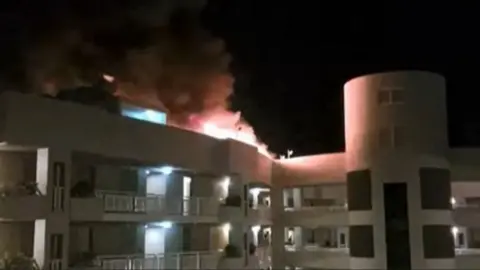 Amanda Kay Flames and smoke pour from the top of a multi-storey white building at night.