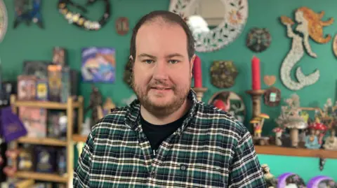 Andy Booth with brown hair and a brown beard. He is wearing a black shirt underneath a green, cream and black checked shirt and standing in front of shelves full of goods.