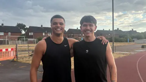 Two men standing on a running track with their arms round each other's shoulders. The man on the left has short black hair and is wearing a black Nike vest. The man on the right has short brown hair and is also wearing a black vest. The sky behind them is cloudy and grey.