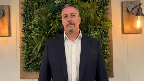 A man with grey hair and a grey and a grey beard he is stood in front of a green floral board