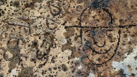 The top of a sandy coloured concrete WW2 anti-tank cube. On the right is a cartoon soldier who is smiling and wearing a helmet.